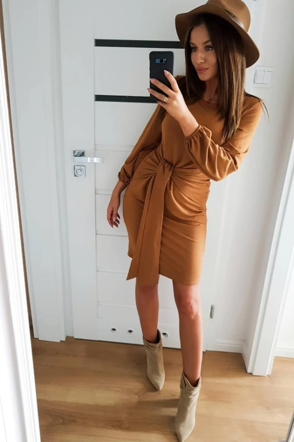Ruda suknelė su ilgu rankoviu ir elegantišku mazgu ties juosmeniu suteikia rafinuotą ir stilingą išvaizdą. Ji puikiai dera su smėlio spalvos skrybėle ir batais, sukuriant harmoningą ir madingą derinį.